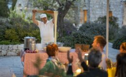 A chef performing a dough stretching demonstration at an outdoor dining event, with guests clapping and enjoying the culinary experience in a picturesque garden setting.