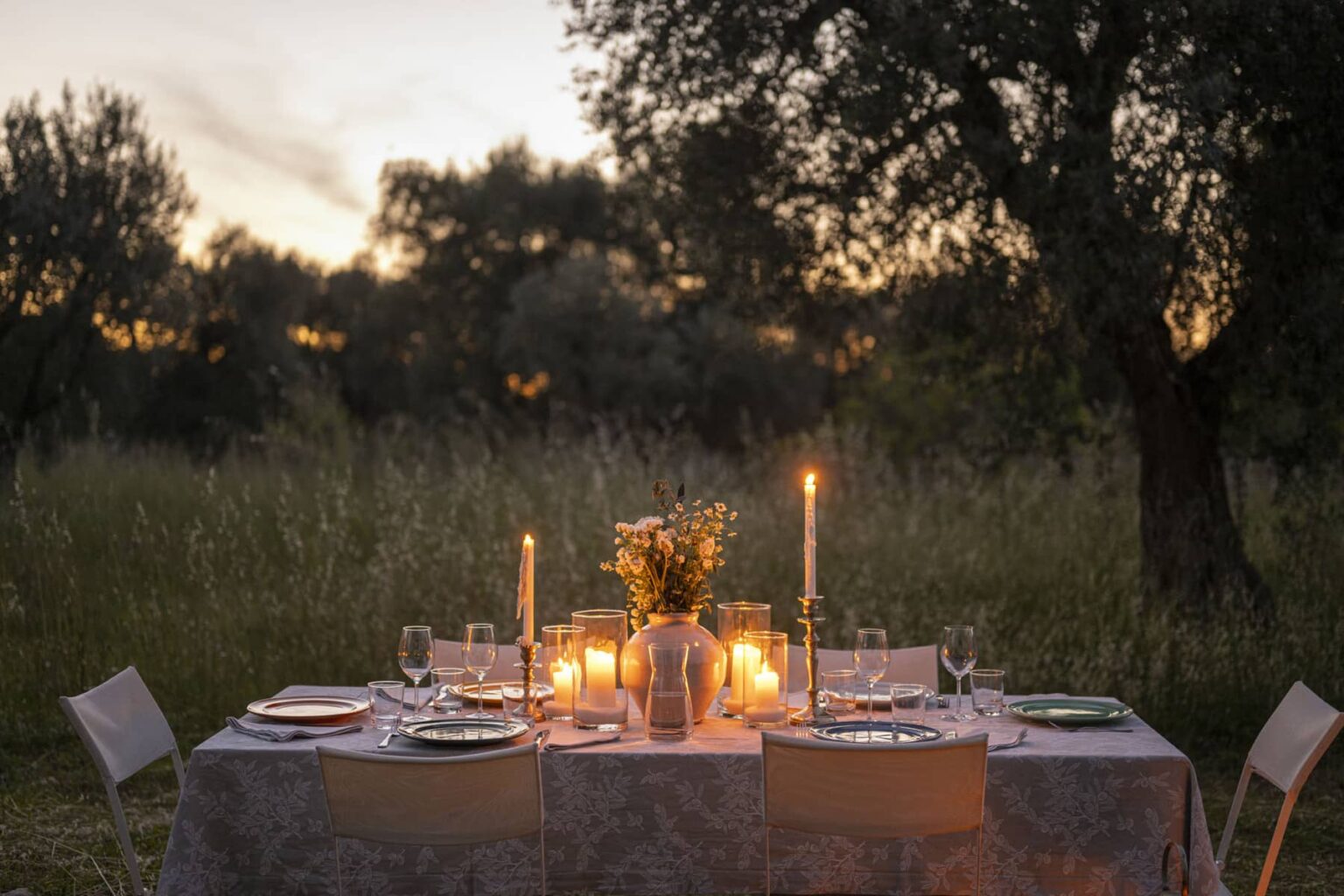 Outdoor dining setup at sunset with a beautifully decorated table, candles, and floral centerpiece surrounded by olive trees. Ideal for intimate gatherings or special occasions. | Only on SopranoVillas