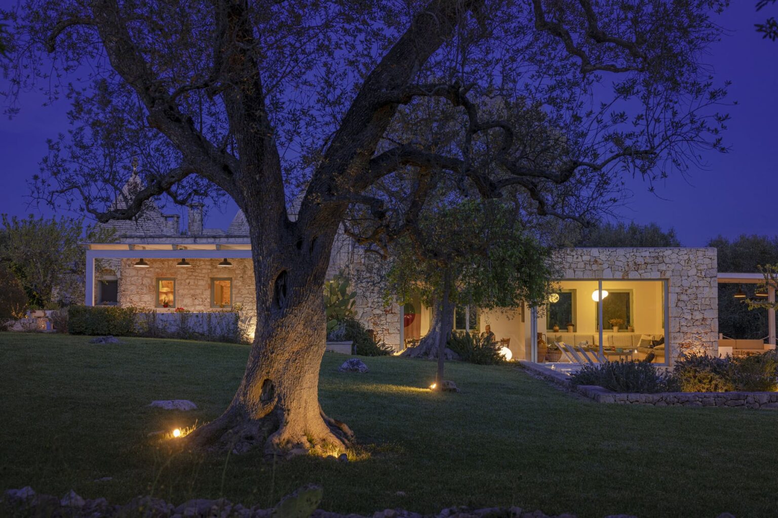 Modern stone house illuminated at night, surrounded by a lush green lawn and a large tree, creating a serene outdoor ambiance. | Only on SopranoVillas