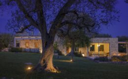 Modern stone house illuminated at night, surrounded by a lush green lawn and a large tree, creating a serene outdoor ambiance.