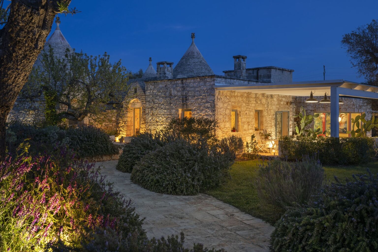 Charming stone villa illuminated at dusk, featuring traditional trulli architecture, surrounded by lush gardens and a winding pathway. | Only on SopranoVillas
