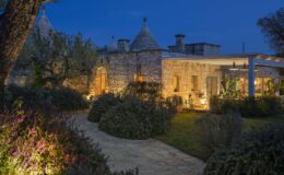 Charming stone villa illuminated at dusk, featuring traditional trulli architecture, surrounded by lush gardens and a winding pathway.
