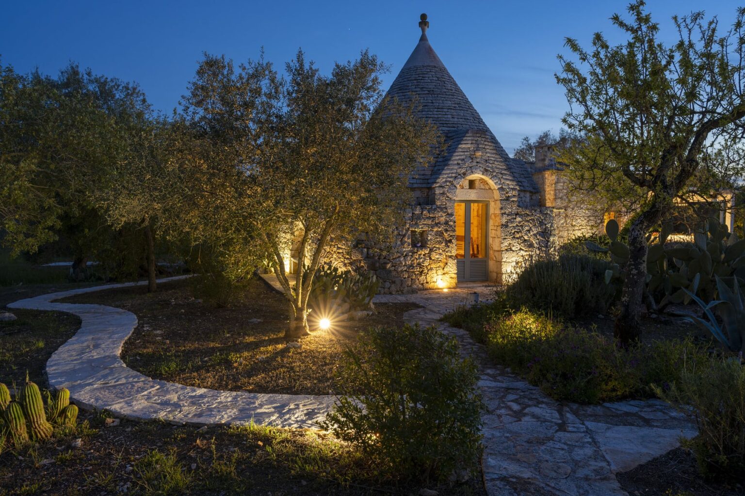Charming stone cottage illuminated at dusk, featuring a winding pathway surrounded by lush greenery and soft lighting, creating an inviting atmosphere for evening strolls. | Only on SopranoVillas