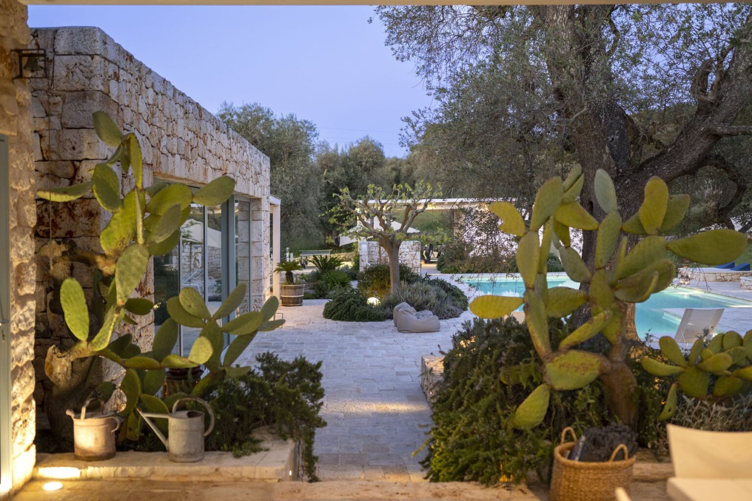 Modern villa exterior with stone walls, lush greenery, and a swimming pool, surrounded by cacti and olive trees, creating a serene outdoor oasis at dusk. | Only on SopranoVillas