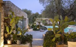 Modern villa exterior with stone walls, lush greenery, and a swimming pool, surrounded by cacti and olive trees, creating a serene outdoor oasis at dusk.