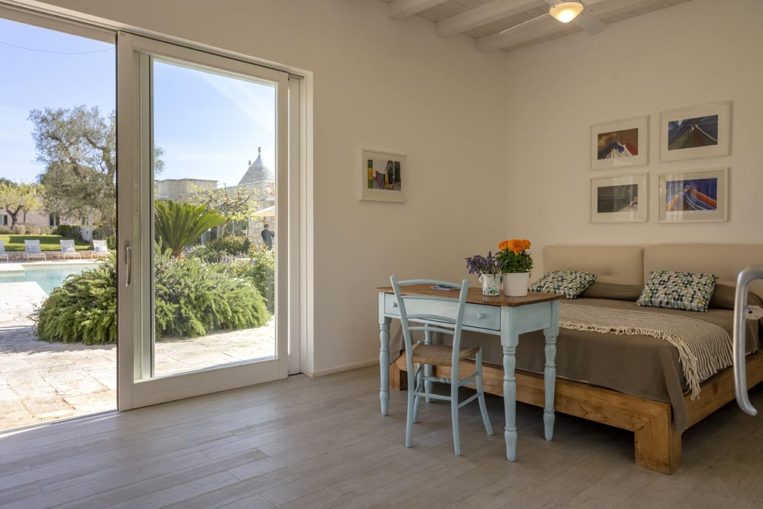 Cozy bedroom interior featuring a wooden bed with decorative pillows, a light blue desk with a chair, and potted flowers. Large glass doors open to a serene outdoor space with a pool and lush greenery, enhancing the inviting atmosphere. | Only on SopranoVillas