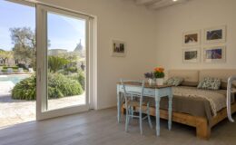 Cozy bedroom interior featuring a wooden bed with decorative pillows, a light blue desk with a chair, and potted flowers. Large glass doors open to a serene outdoor space with a pool and lush greenery, enhancing the inviting atmosphere.