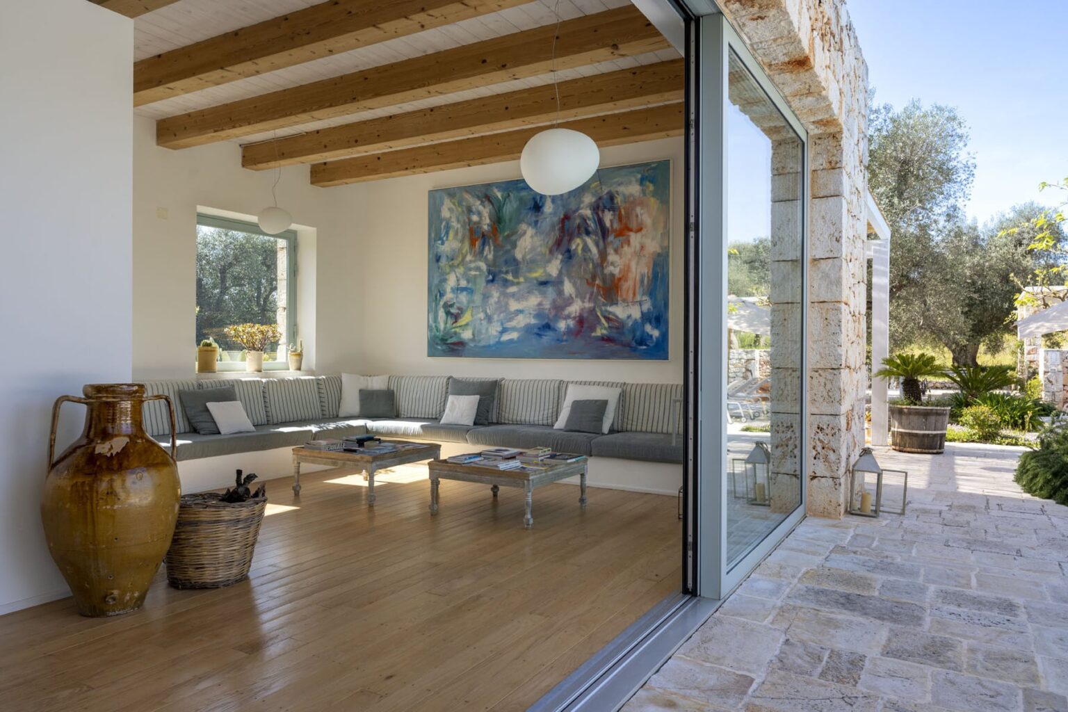 Modern living room with large glass doors opening to an outdoor space, featuring wooden beams, a colorful artwork on the wall, and stylish furniture. | Only on SopranoVillas