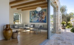 Modern living room with large glass doors opening to an outdoor space, featuring wooden beams, a colorful artwork on the wall, and stylish furniture.