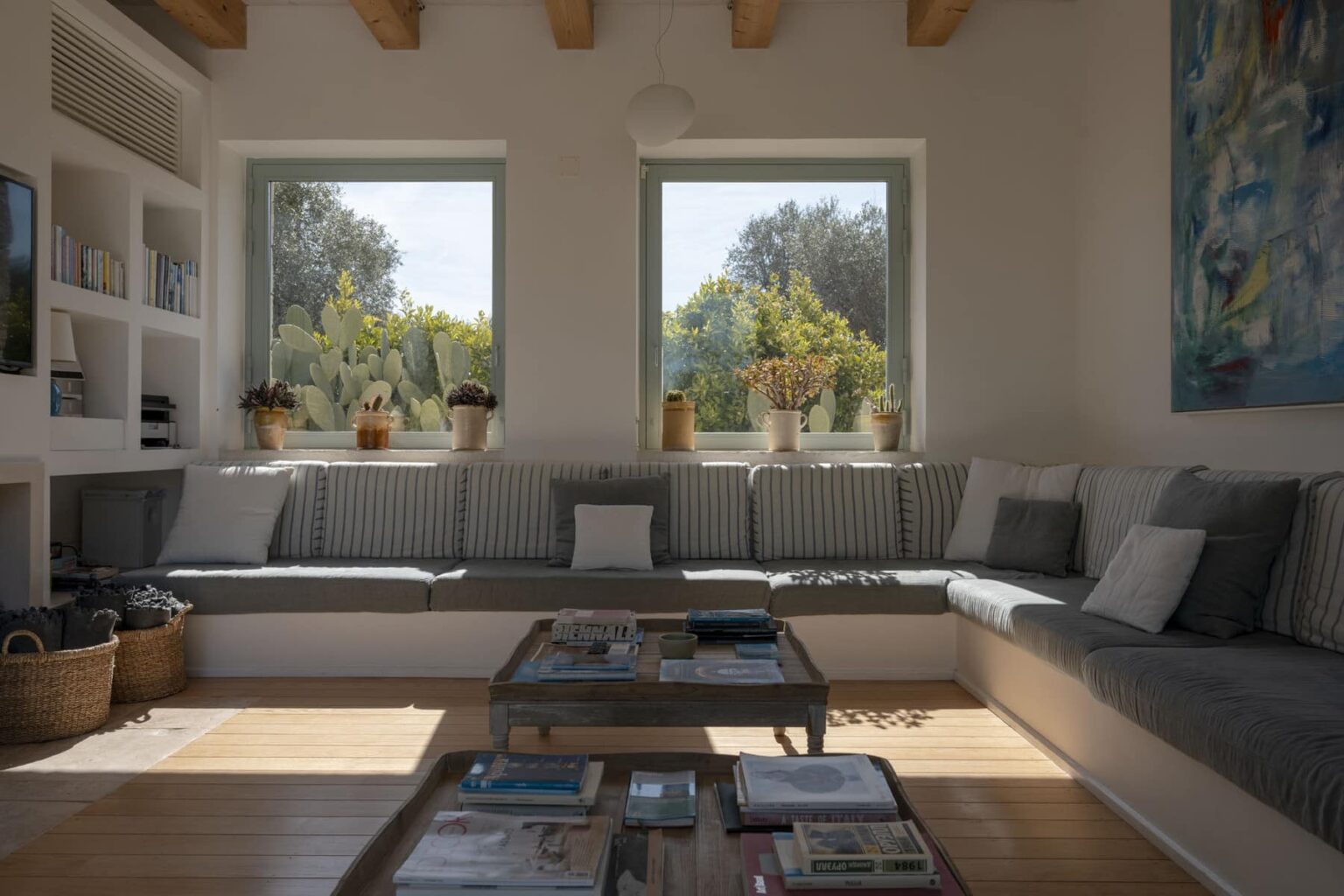 Cozy living room featuring a spacious sectional sofa with striped cushions, natural light streaming through large windows, and a wooden coffee table adorned with books and decor. Greenery visible outside enhances the serene atmosphere. | Only on SopranoVillas