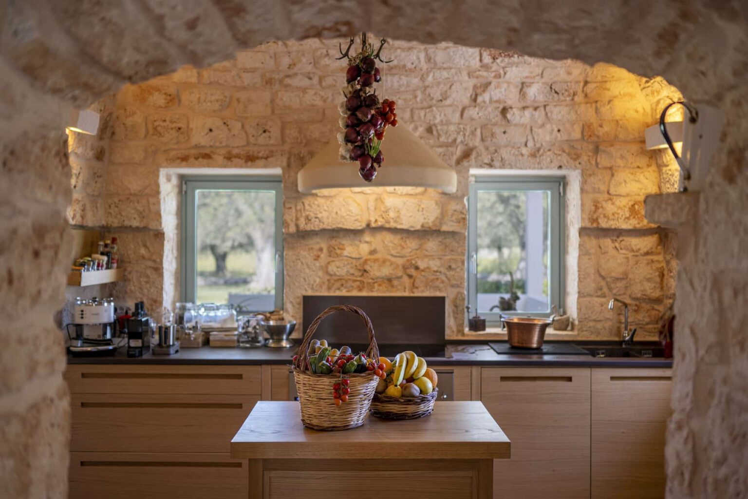 Rustic kitchen interior featuring stone walls, wooden cabinetry, and a central island with two baskets of fresh fruits, including bananas and vegetables, illuminated by natural light from windows. | Only on SopranoVillas