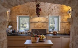 Rustic kitchen interior featuring stone walls, wooden cabinetry, and a central island with two baskets of fresh fruits, including bananas and vegetables, illuminated by natural light from windows.