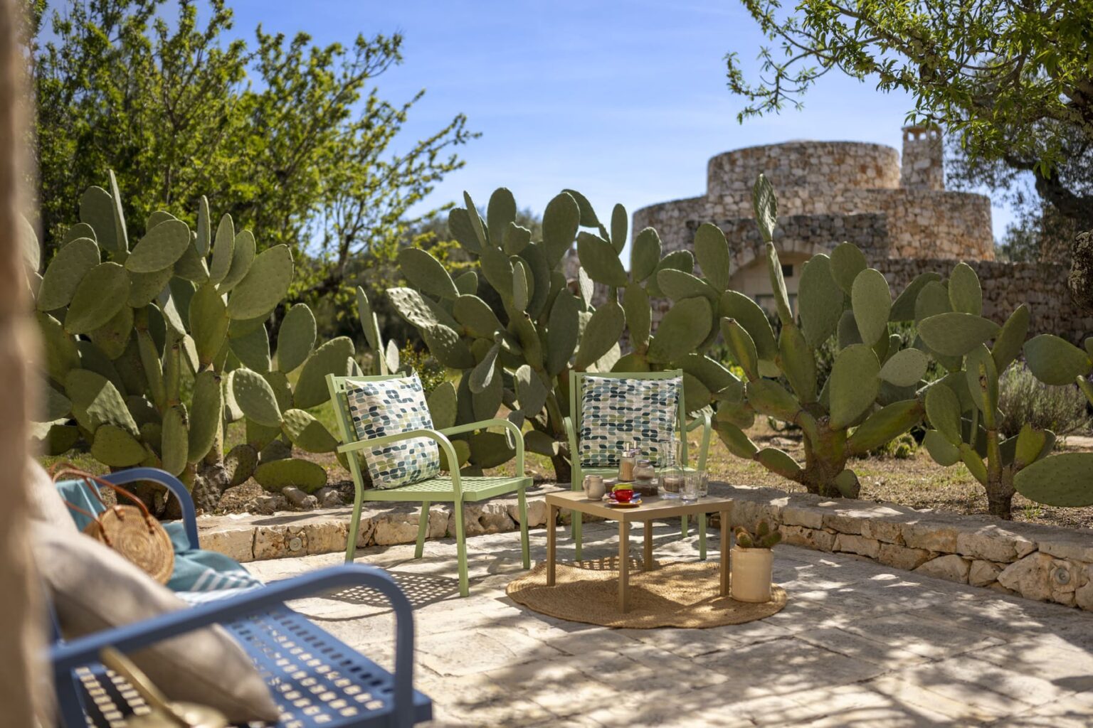 Cozy outdoor seating area with patterned cushions, surrounded by cacti and greenery, featuring a small table and a historic stone structure in the background under a clear blue sky. | Only on SopranoVillas