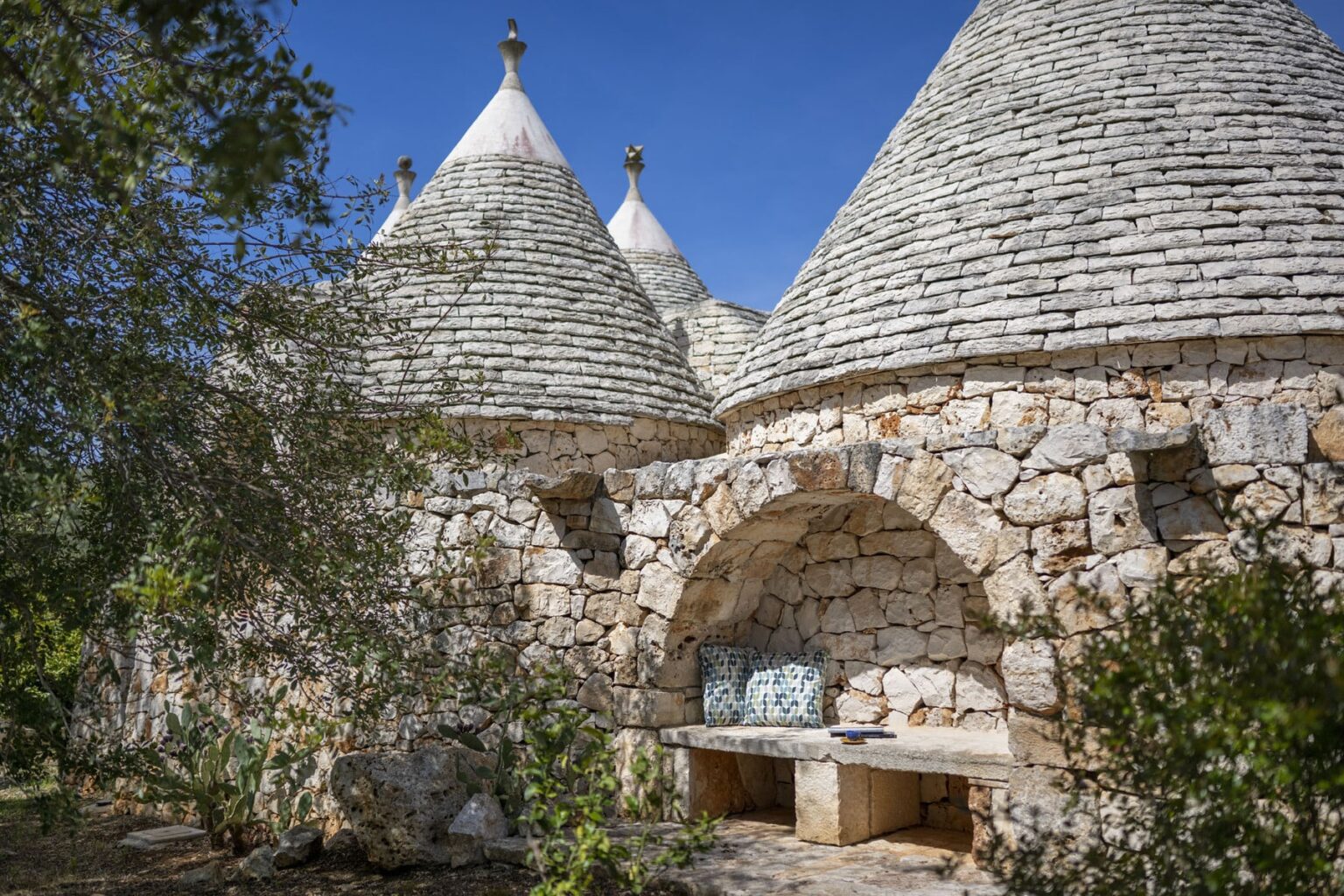 Trullo houses with conical roofs made of stone, surrounded by greenery and a rustic stone bench, set against a clear blue sky. | Only on SopranoVillas
