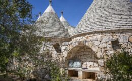 Trullo houses with conical roofs made of stone, surrounded by greenery and a rustic stone bench, set against a clear blue sky.