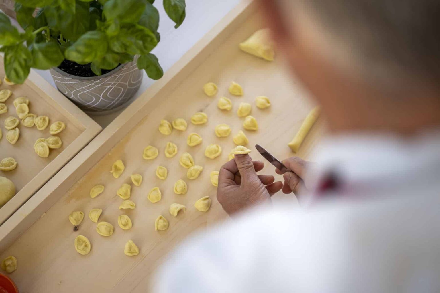 A person shaping fresh pasta on a wooden surface, surrounded by uncooked pasta pieces and a potted plant in the background. | Only on SopranoVillas