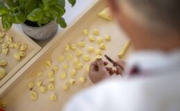 A person shaping fresh pasta on a wooden surface, surrounded by uncooked pasta pieces and a potted plant in the background.