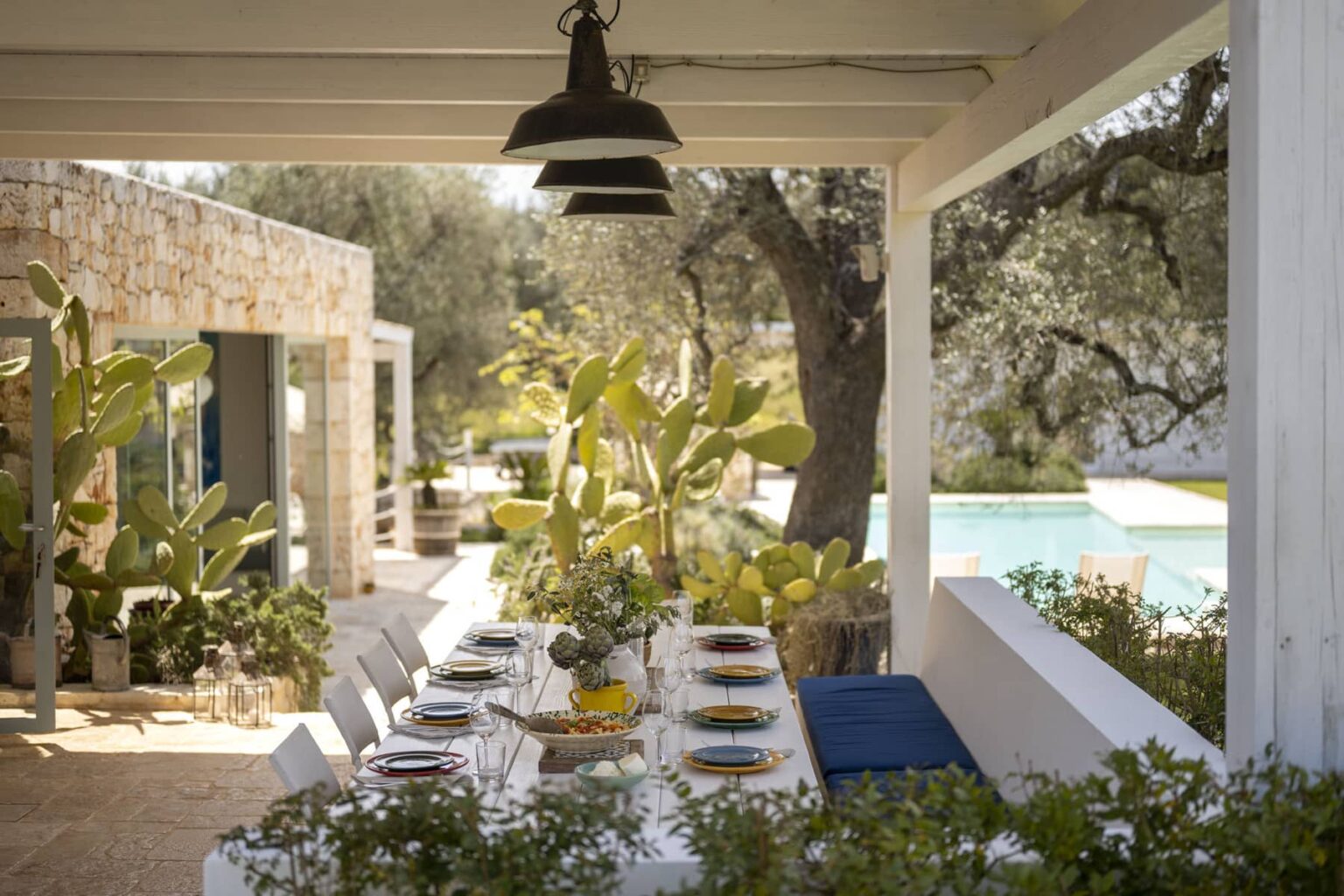 Outdoor dining area featuring a long table set with colorful plates and glassware, surrounded by lush greenery and cacti, with a view of a swimming pool in the background. The space is bathed in natural light, enhancing the inviting atmosphere for gatherings. | Only on SopranoVillas