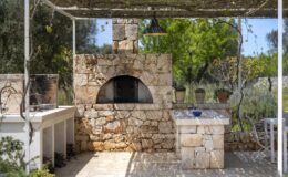 Outdoor stone kitchen featuring a traditional wood-fired oven, a spacious countertop, and surrounding greenery, ideal for grilling and entertaining.