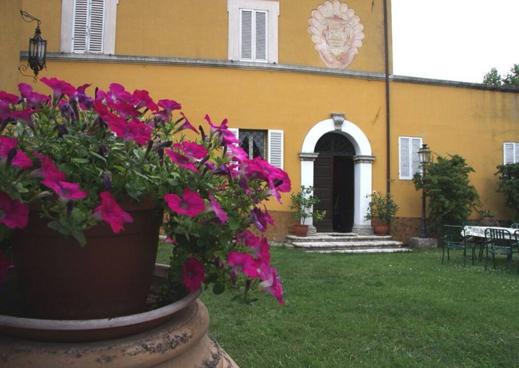 Colorful pink petunias in a terracotta pot in the foreground, with a charming yellow building featuring white window shutters and an arched entrance in the background, set in a lush green garden. | Only on SopranoVillas