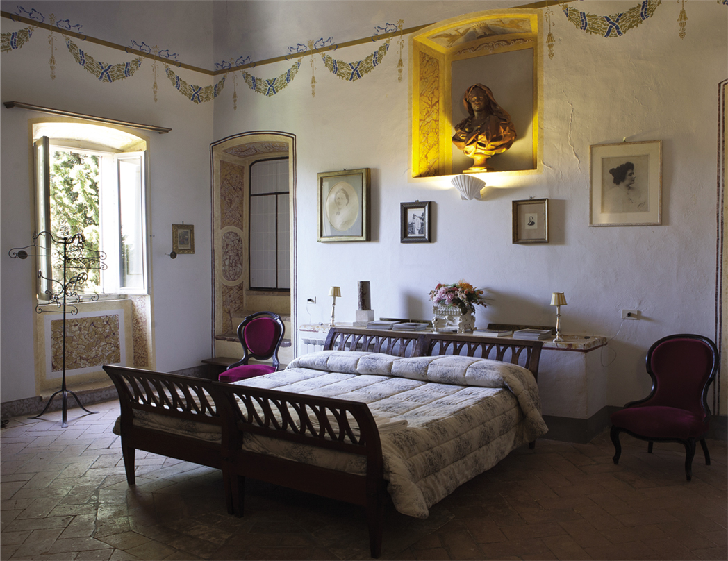 Elegant bedroom interior featuring a wooden bed with a quilt, two vintage chairs, and decorative wall art. Natural light floods the room through large windows, enhancing the serene atmosphere. Ideal for showcasing home decor and design inspiration. | Only on SopranoVillas