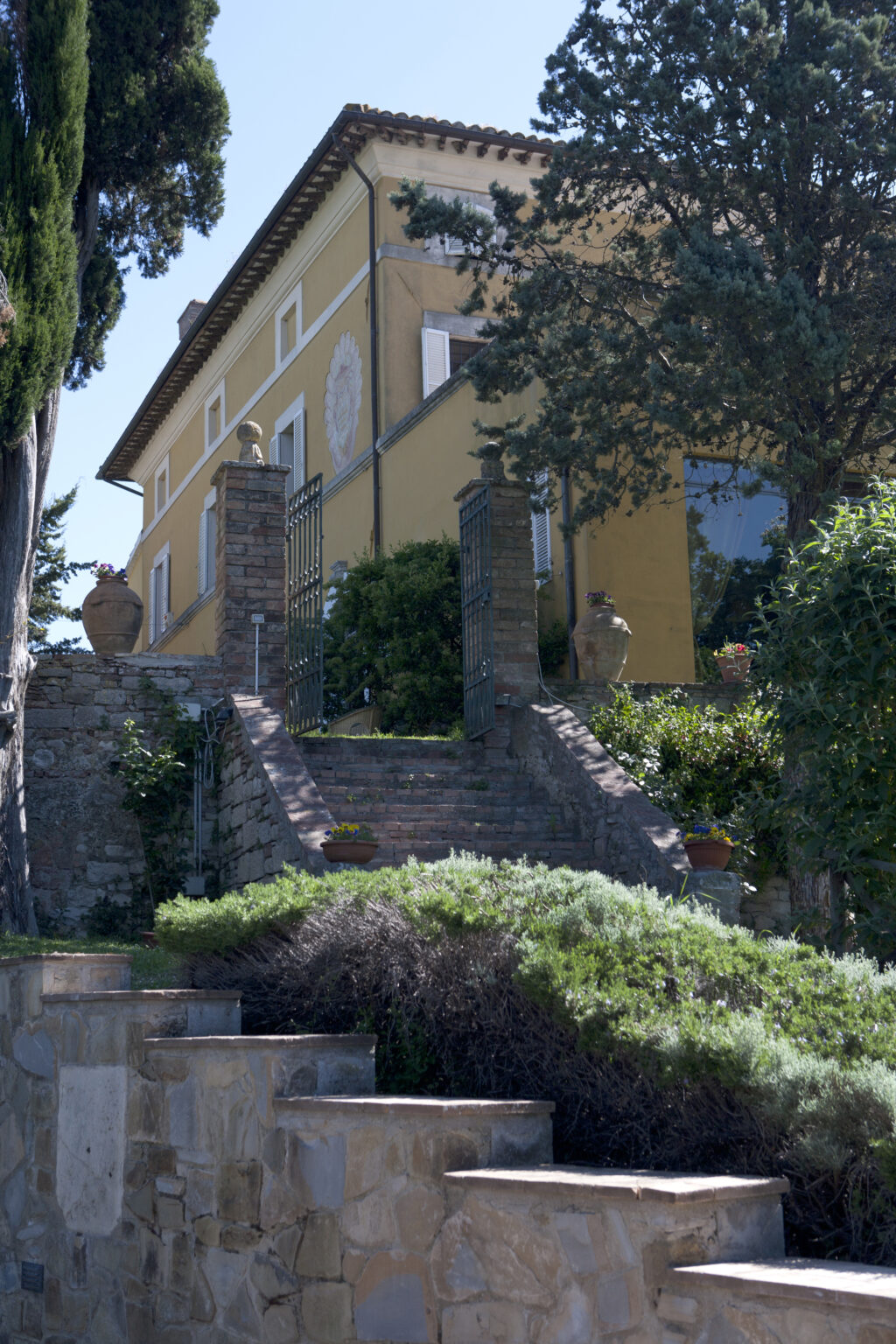 Historic yellow villa with stone steps and wrought iron gates, surrounded by lush greenery and potted plants, set against a clear blue sky. | Only on SopranoVillas