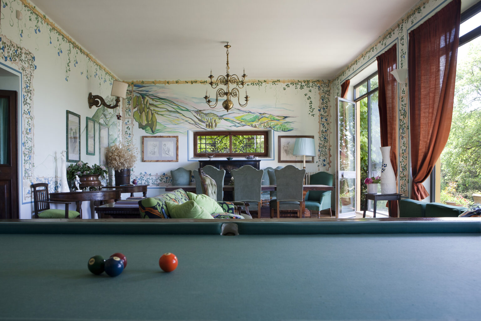 Elegant interior of a spacious game room featuring a green billiard table, decorative floral wallpaper, a chandelier, and a view of the garden through large windows. | Only on SopranoVillas