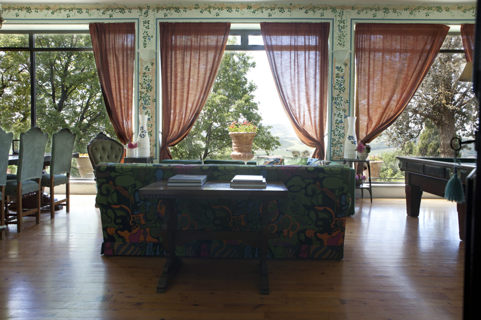 Cozy living room with a vibrant patterned sofa, large windows adorned with sheer curtains, and a scenic view of greenery outside. A wooden table sits in the foreground, enhancing the inviting atmosphere of the space. | Only on SopranoVillas