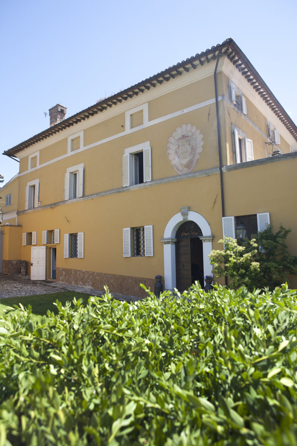 Exterior view of a yellow building featuring white shutters, arched doorway, and decorative wall artwork, surrounded by greenery. Ideal for showcasing architectural design and landscaping. | Only on SopranoVillas