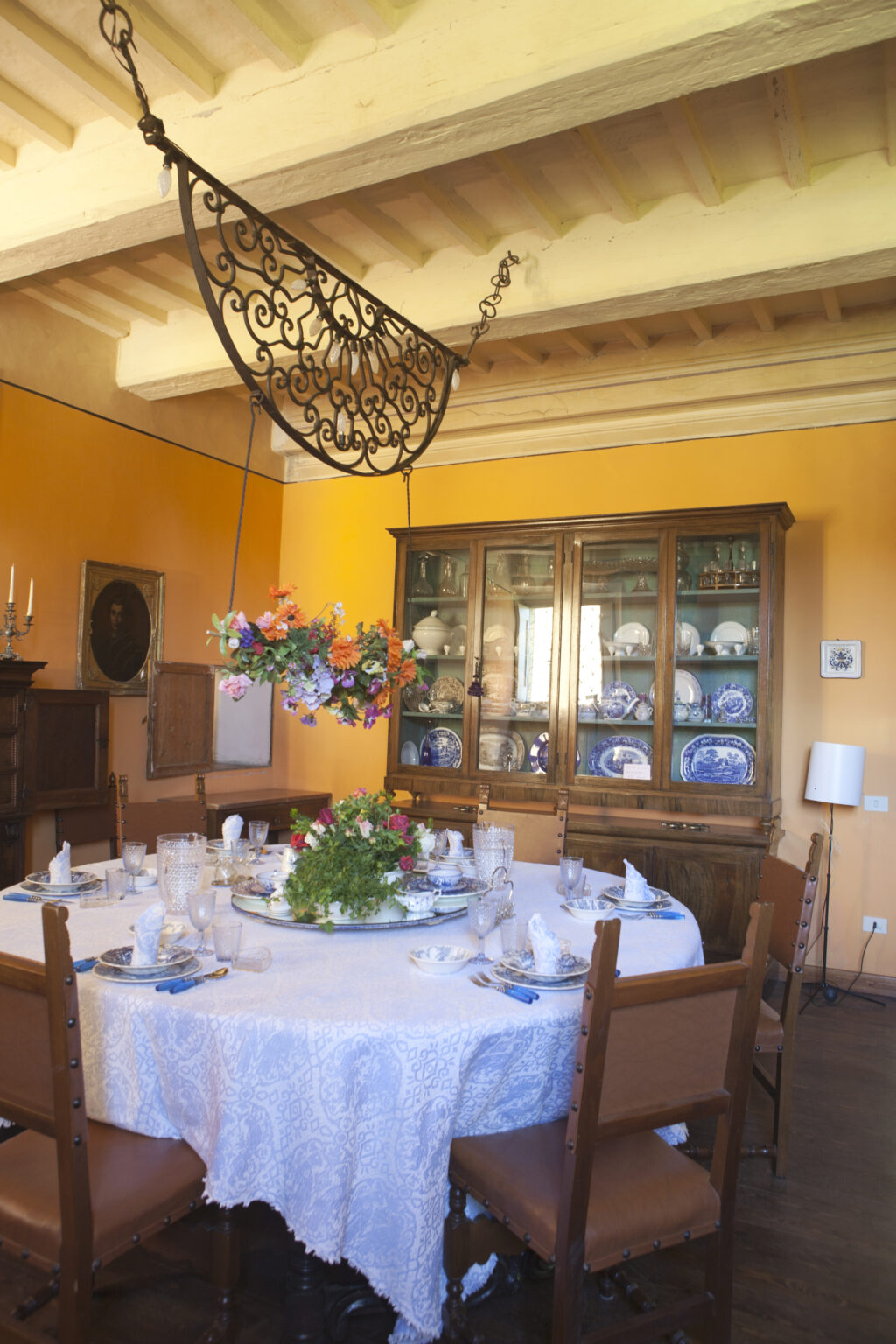 Elegant dining room featuring a round table set for a meal, adorned with white tableware and a floral centerpiece. The walls are painted a warm orange, and a wooden cabinet displays fine china and glassware. A decorative chandelier hangs from the ceiling, adding charm to the inviting atmosphere. | Only on SopranoVillas