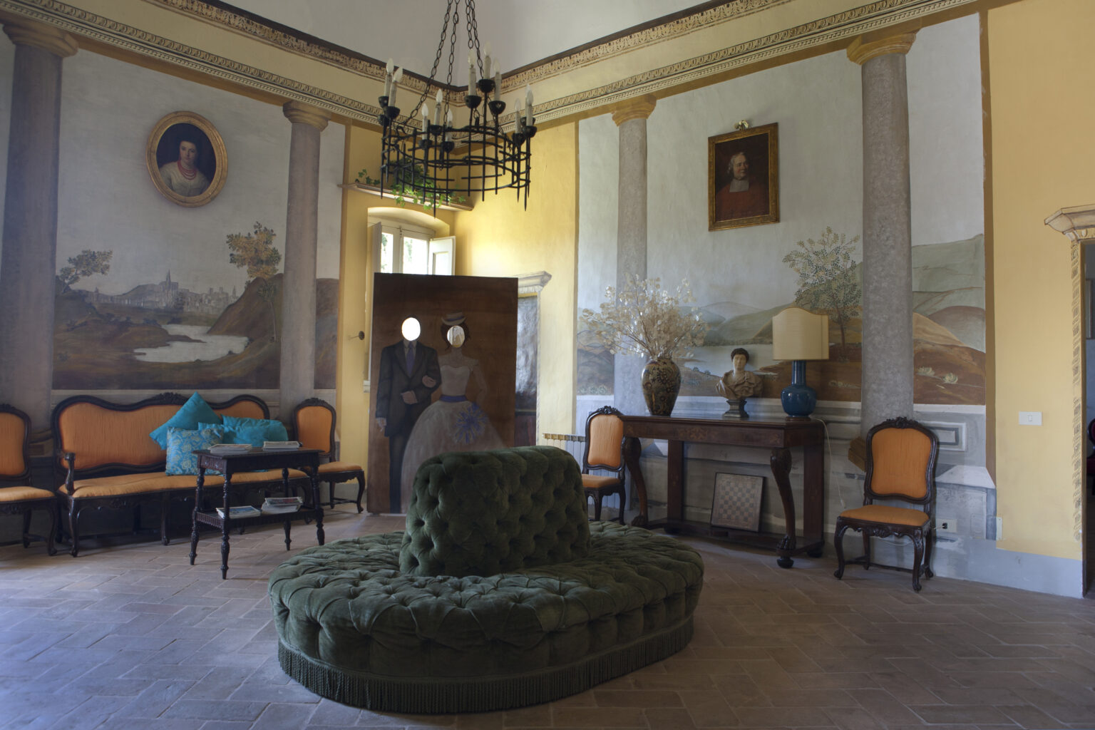 Interior view of a vintage room featuring a green velvet couch, antique furniture, and decorative murals on the walls, illuminated by natural light from large windows. | Only on SopranoVillas
