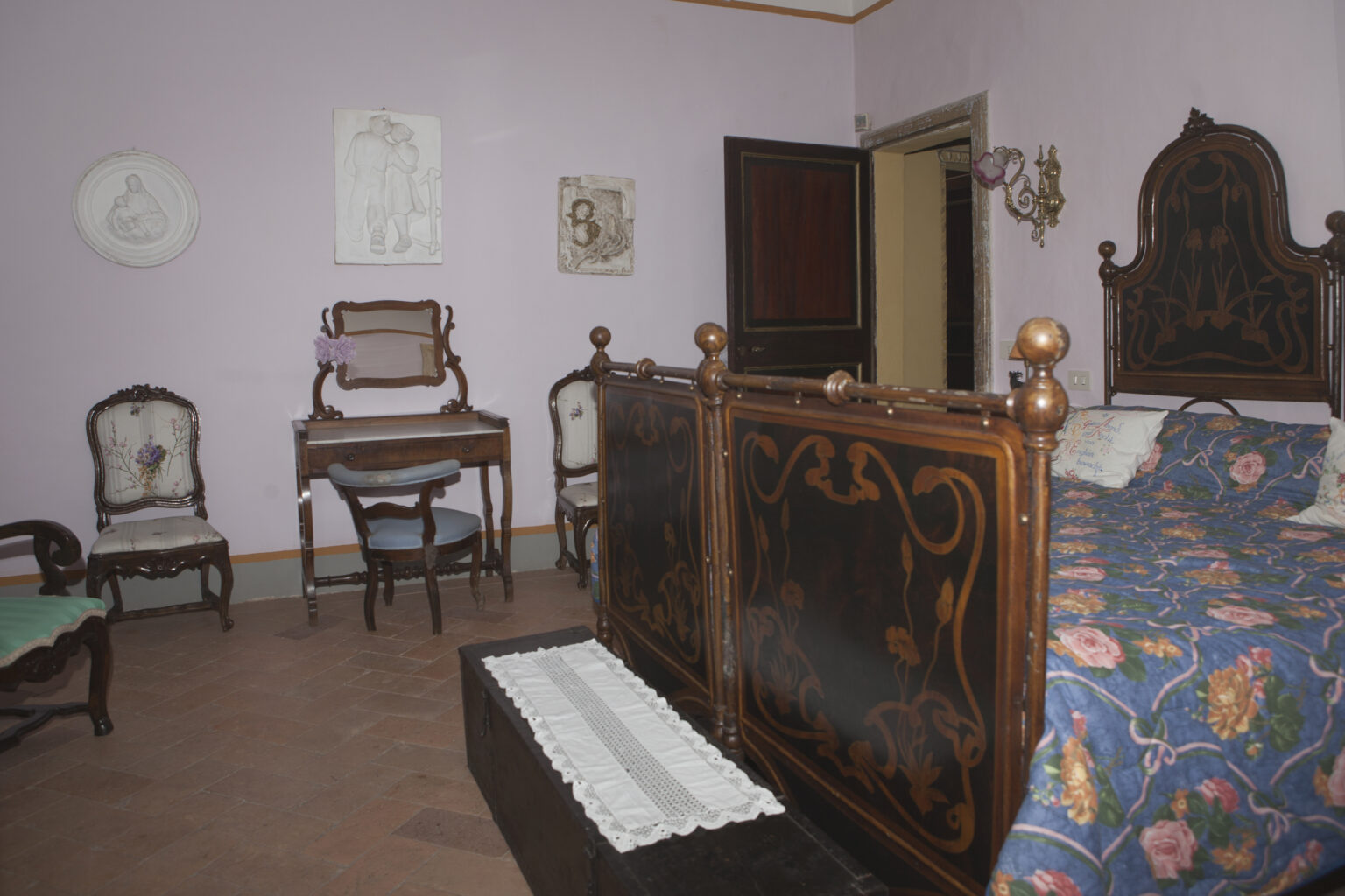 Historic bedroom featuring antique furniture, including a wooden bed with intricate carvings, a decorative quilt, and vintage chairs against a pastel purple wall. The room includes a small writing desk and an open door leading to another area. | Only on SopranoVillas