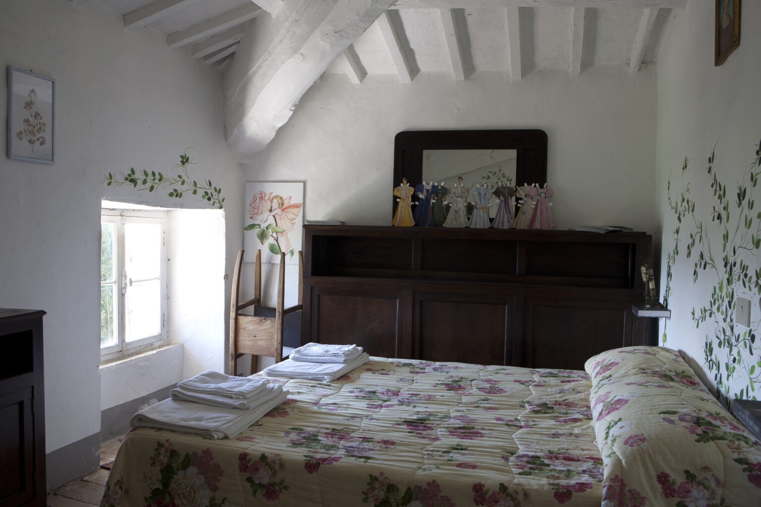 Cozy bedroom featuring a floral-patterned bedspread, wooden furniture, and natural light from a window, with decorative wall art and a mirror reflecting a serene ambiance. | Only on SopranoVillas
