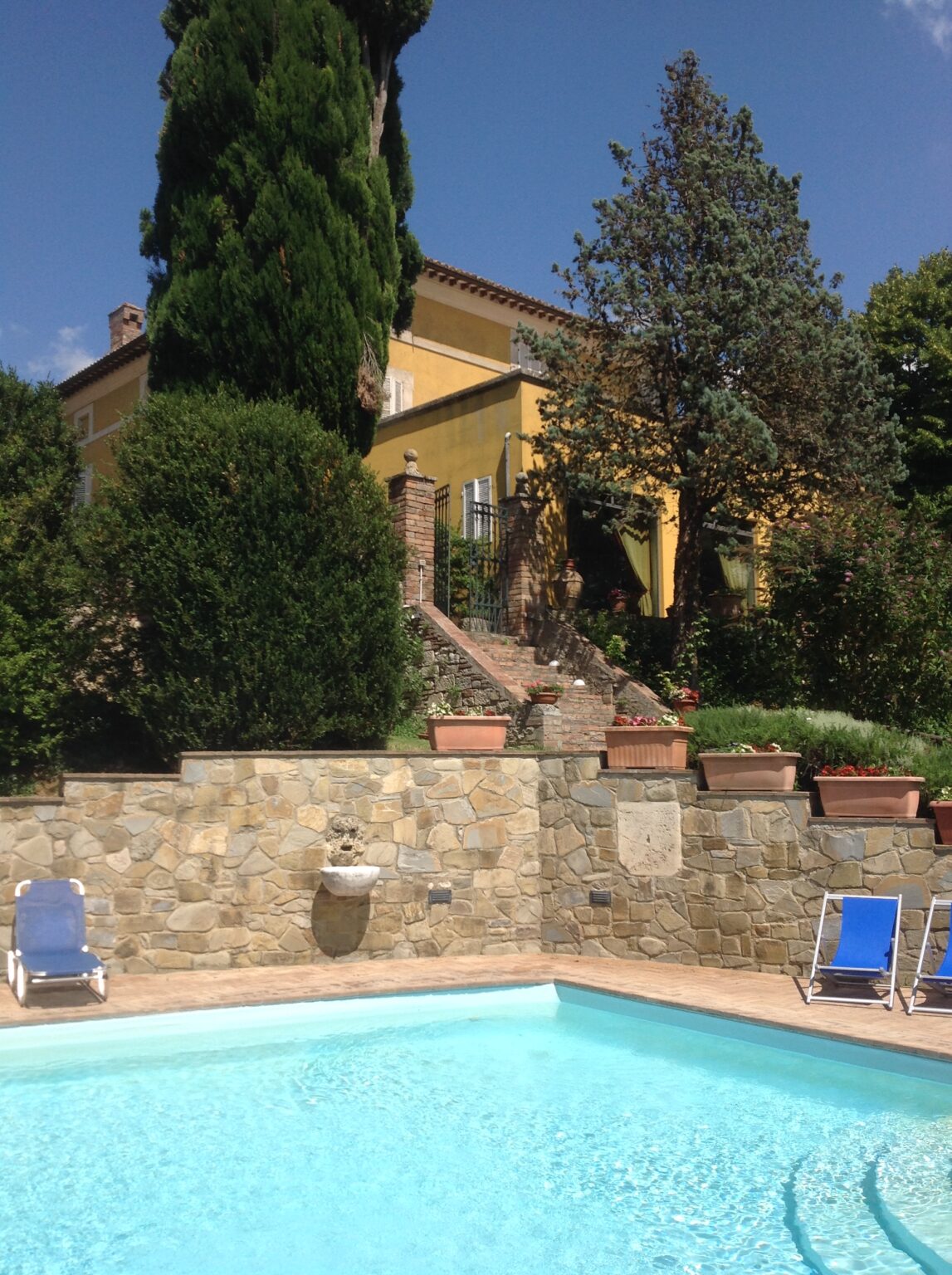 Scenic view of a yellow villa surrounded by lush greenery and stone walls, featuring a staircase leading to the entrance and potted plants lining the pathway. | Only on SopranoVillas