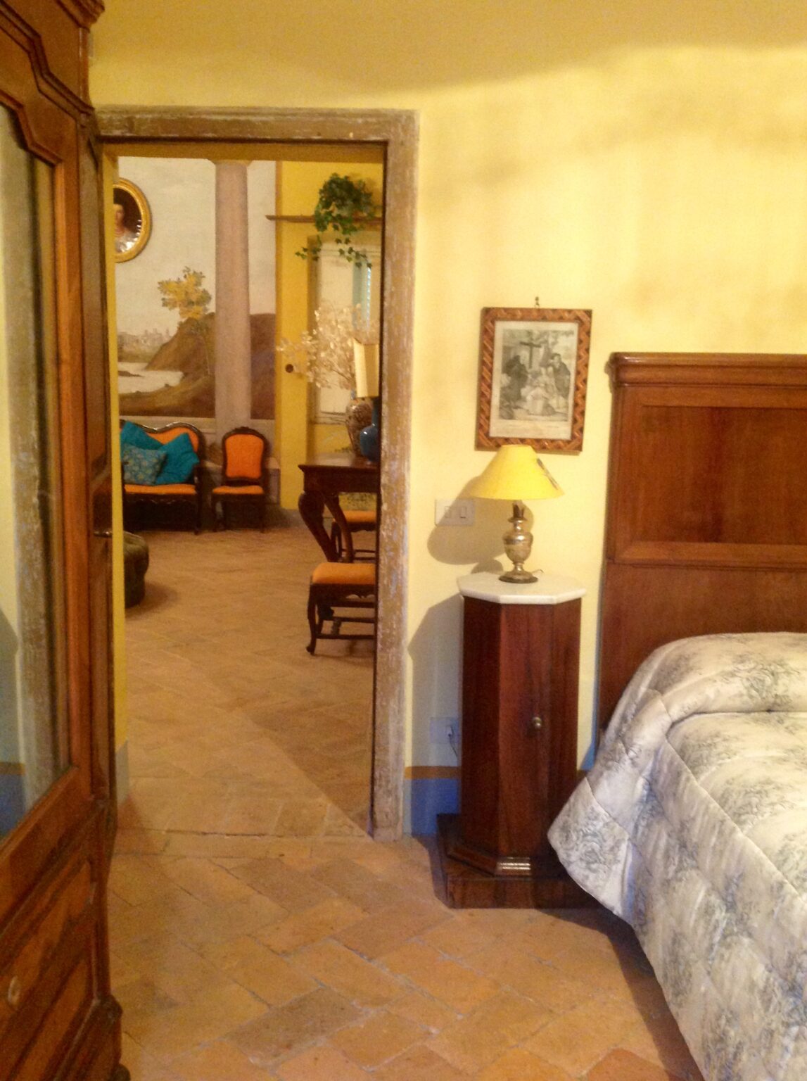 Cozy bedroom with a wooden bed and nightstand, featuring a yellow lamp. A doorway leads to a living area with vintage furniture and a mural, showcasing a warm, inviting atmosphere. The room has terracotta tiled flooring and soft yellow walls. | Only on SopranoVillas