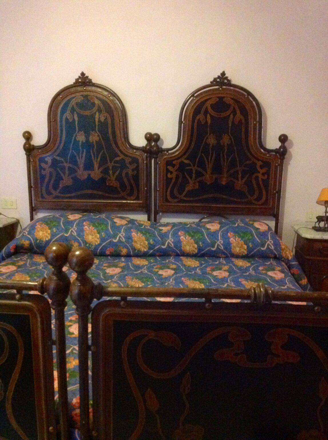 Antique wooden bed with intricately designed headboards featuring floral patterns, complemented by a colorful floral bedspread. The cozy bedroom setting includes a nightstand with a lamp, enhancing the vintage aesthetic. | Only on SopranoVillas