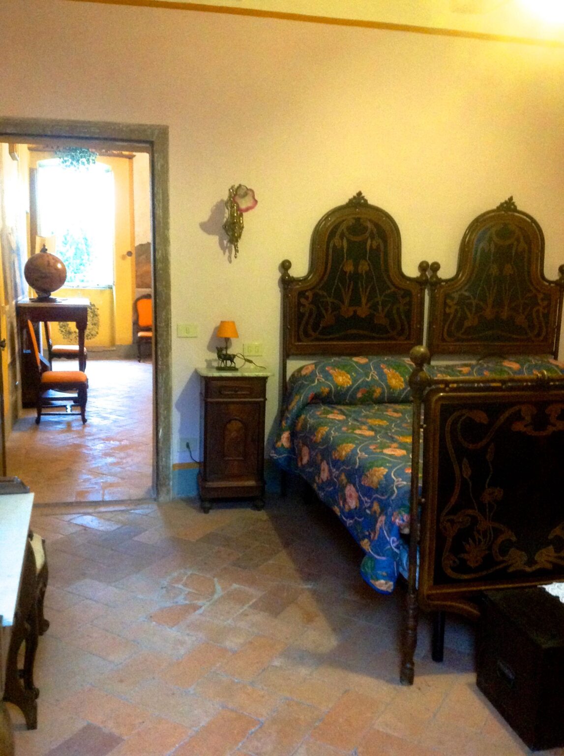 Cozy vintage bedroom featuring a double bed with an intricately designed headboard, floral bedding, a bedside table with a lamp, and a glimpse of a well-furnished adjacent room, highlighting rustic charm and inviting atmosphere. | Only on SopranoVillas