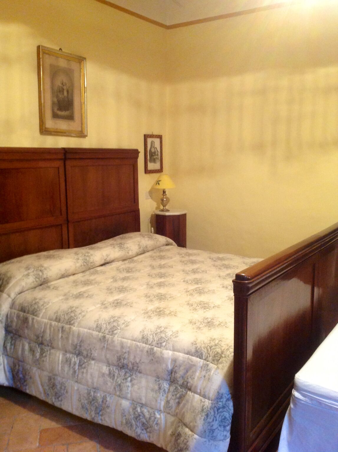 Cozy bedroom featuring a wooden bed frame with a floral-patterned comforter, a bedside lamp, and warm yellow walls, creating a welcoming atmosphere for relaxation. | Only on SopranoVillas
