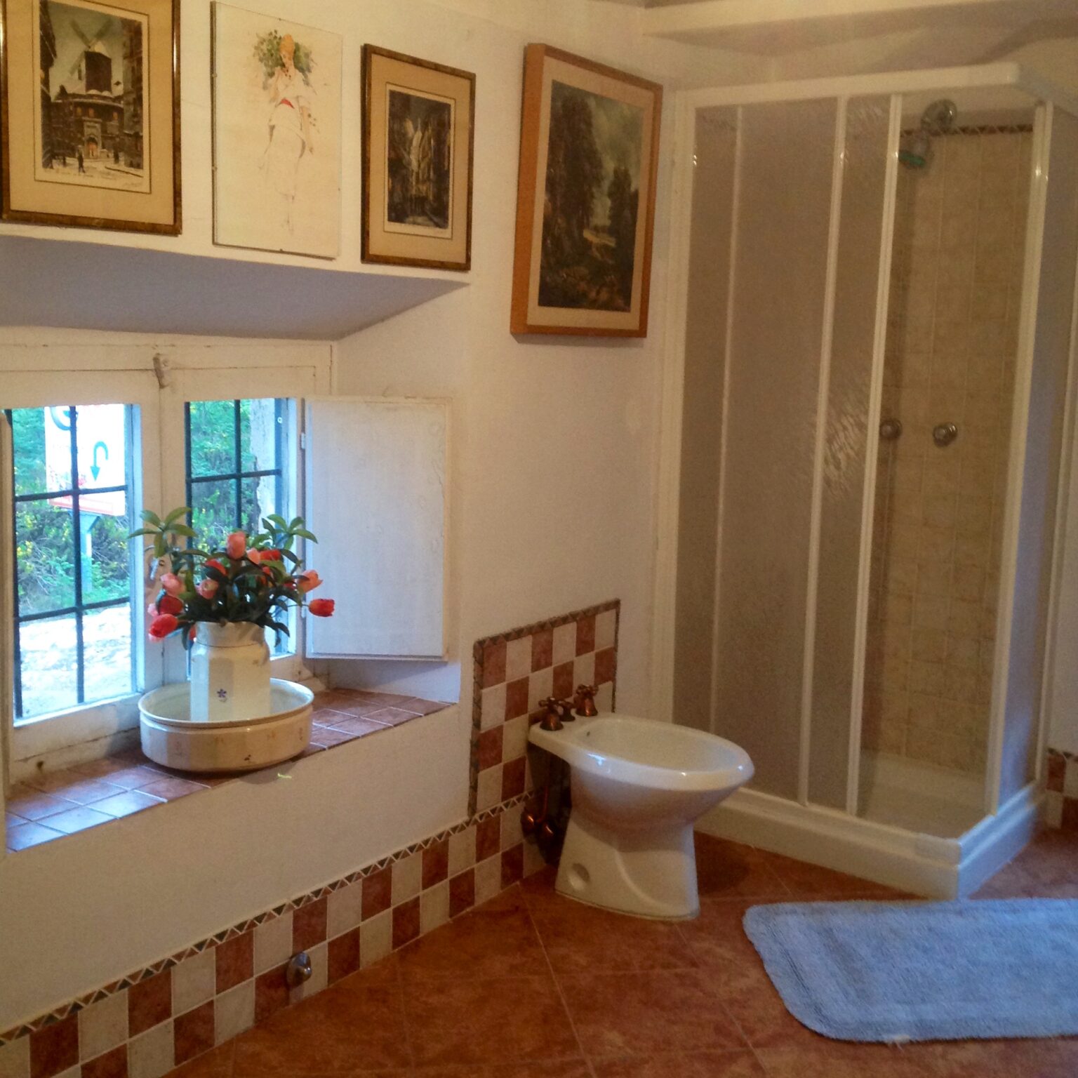 Cozy bathroom interior featuring a white toilet, a glass shower enclosure, and a window with decorative flowers. The walls are adorned with framed pictures, and the tiled floor adds a rustic charm. | Only on SopranoVillas