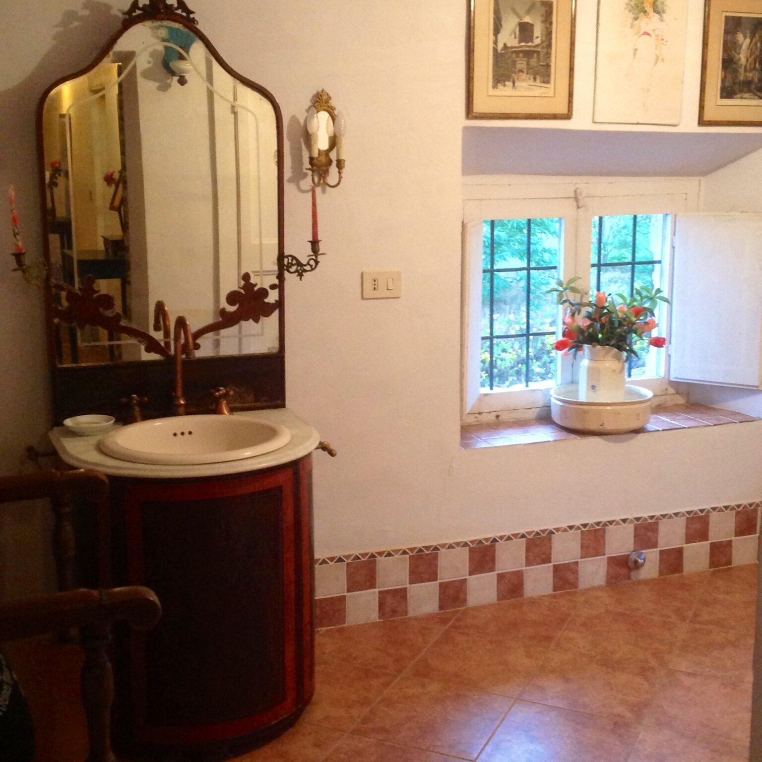 Vintage bathroom interior featuring a decorative sink with a mirror, a window with floral decor, and checkered tile flooring. Natural light enhances the cozy atmosphere. | Only on SopranoVillas