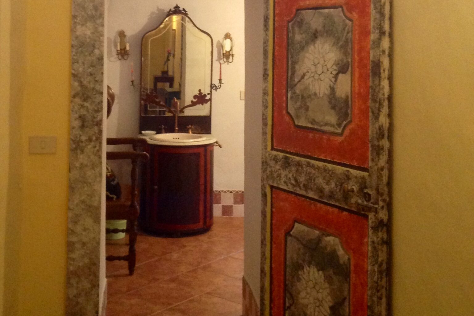 Interior view of a decorative bathroom with a vintage mirror and ornate red door, showcasing rustic tiles and warm lighting. | Only on SopranoVillas