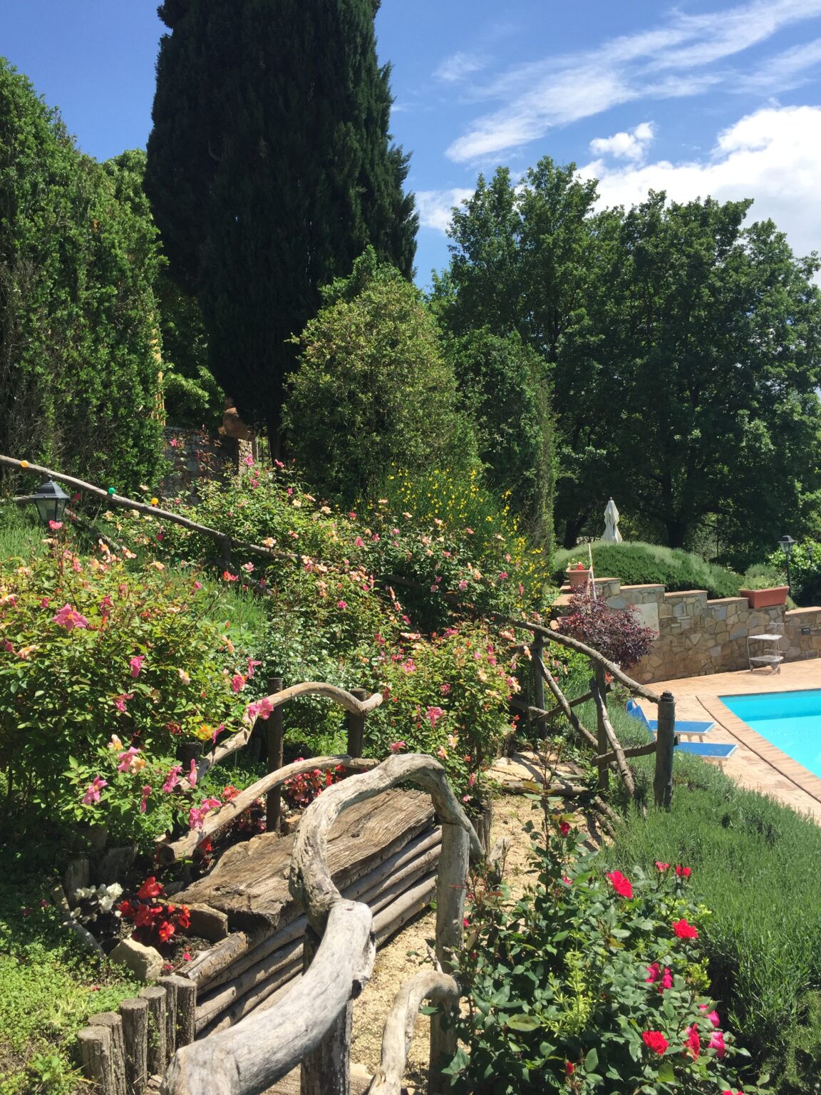 Lush garden with vibrant flowers and greenery surrounding a tranquil swimming pool under a clear blue sky. Features rustic wooden fencing and a variety of plants, enhancing the serene outdoor atmosphere. | Only on SopranoVillas