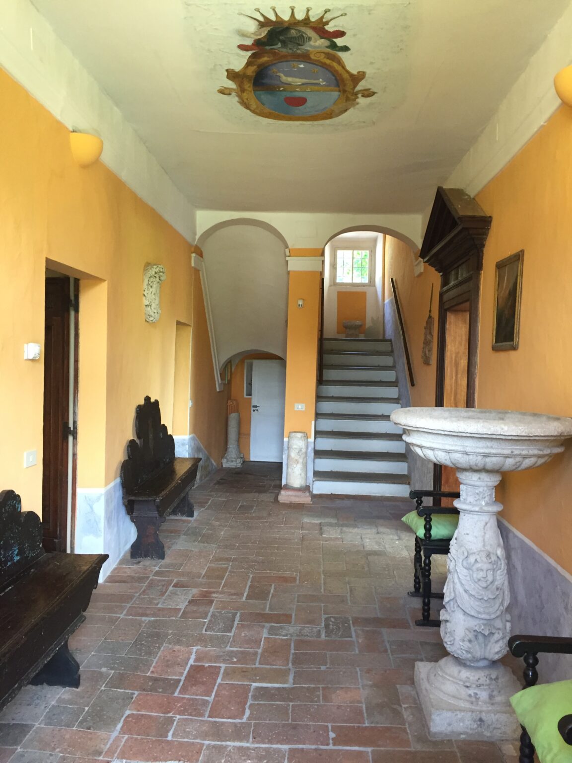 Interior view of a spacious hallway featuring warm orange walls, a staircase leading to the upper floor, and a decorative stone basin. The hallway includes wooden benches and artwork, creating an inviting atmosphere. Natural light filters in through a window at the top of the stairs. | Only on SopranoVillas