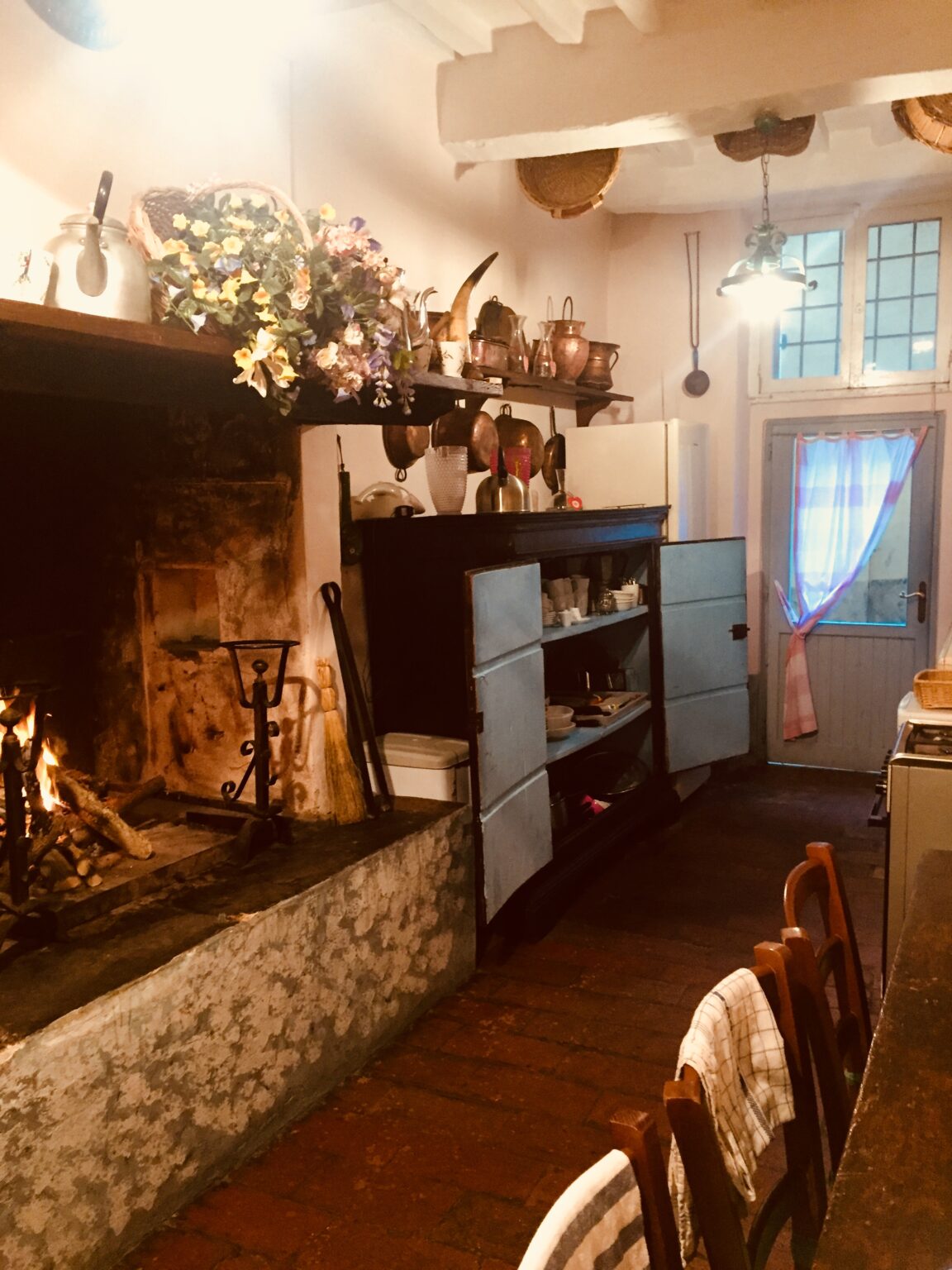 Cozy kitchen interior featuring a traditional stone fireplace, vintage wooden cabinets, and a charming door with a light curtain. The space is adorned with rustic kitchenware and a warm, inviting atmosphere. | Only on SopranoVillas