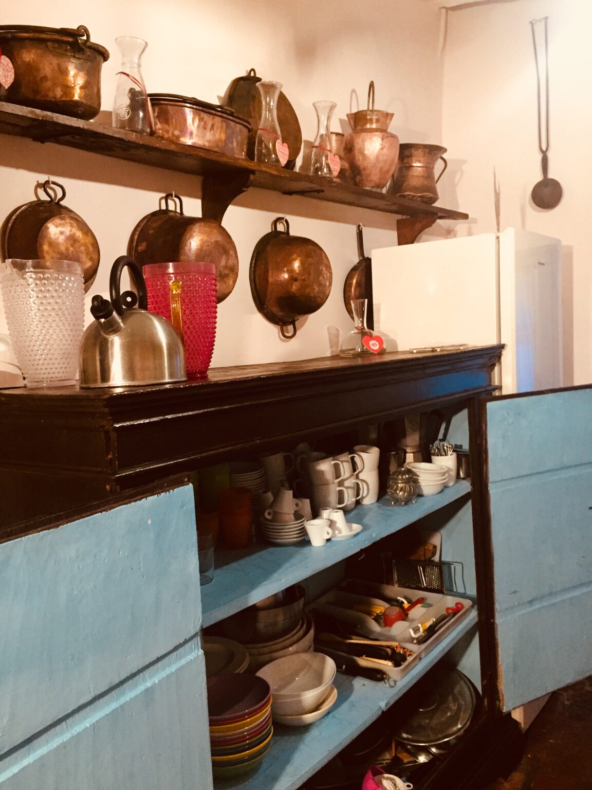 A rustic kitchen interior featuring a wooden cabinet with blue doors, displaying various dishes and mugs. Above, there are copper pots hanging on the wall, alongside a kettle and colorful glassware. A white refrigerator is visible in the background, adding to the cozy atmosphere. | Only on SopranoVillas