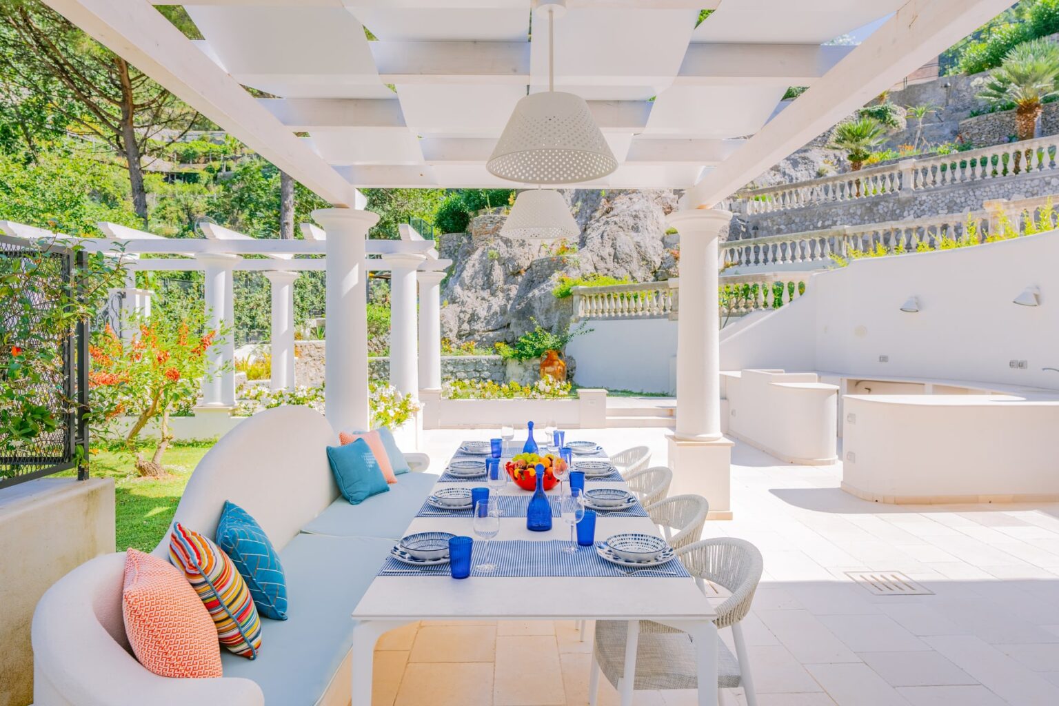 Spacious outdoor dining area featuring a long table set with blue glasses and decorative plates, surrounded by lush greenery and elegant white columns. The area is bright and airy, perfect for al fresco meals, with colorful cushions on a nearby sofa and a backdrop of natural stone and landscaped terraces. | Only on SopranoVillas