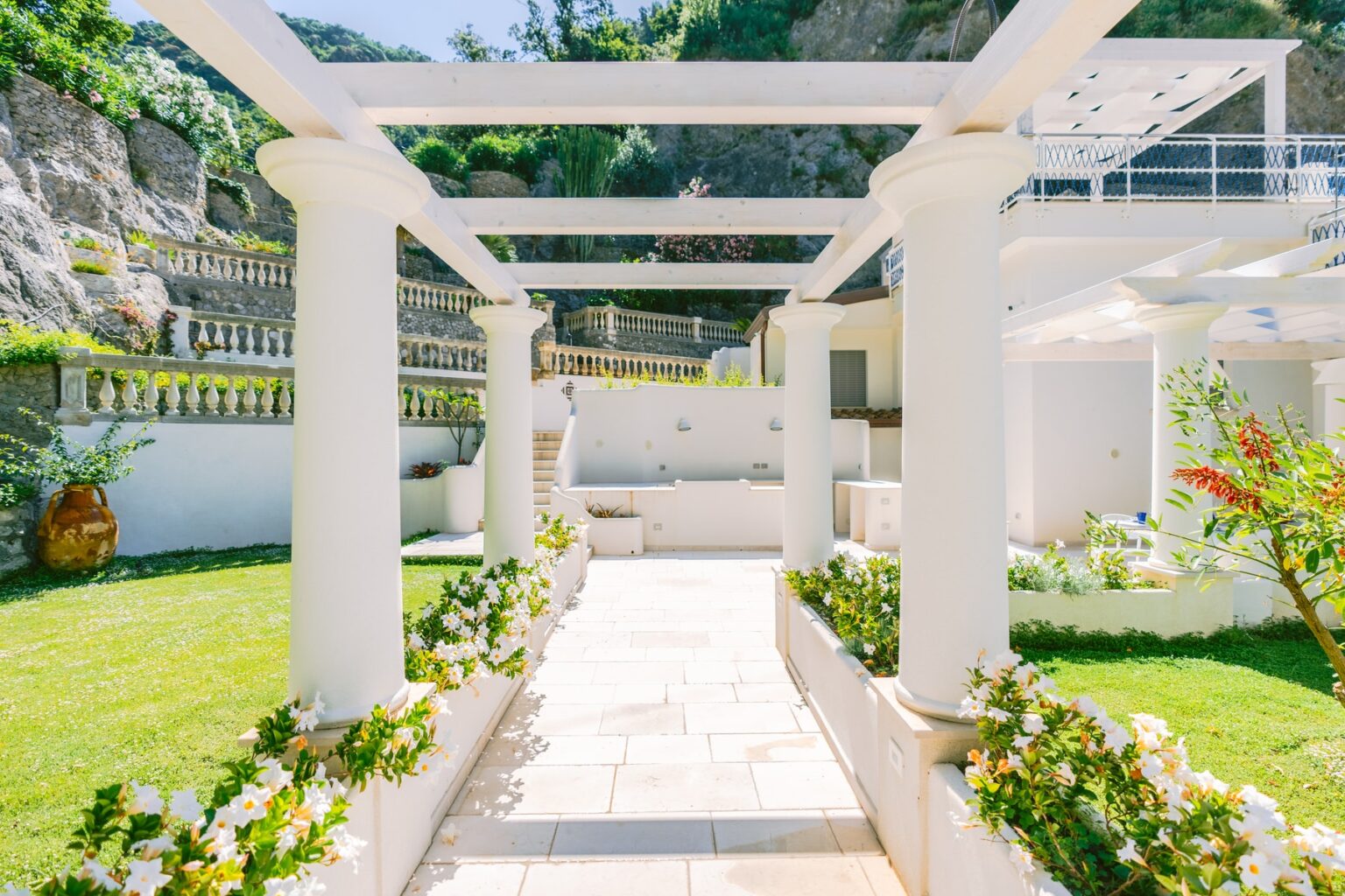 Beautiful outdoor pathway leading through white columns and lush greenery, featuring blooming flowers and a well-maintained garden, set against a scenic backdrop of rocky hills and blue skies. Ideal for showcasing elegant landscaping and outdoor living spaces. | Only on SopranoVillas