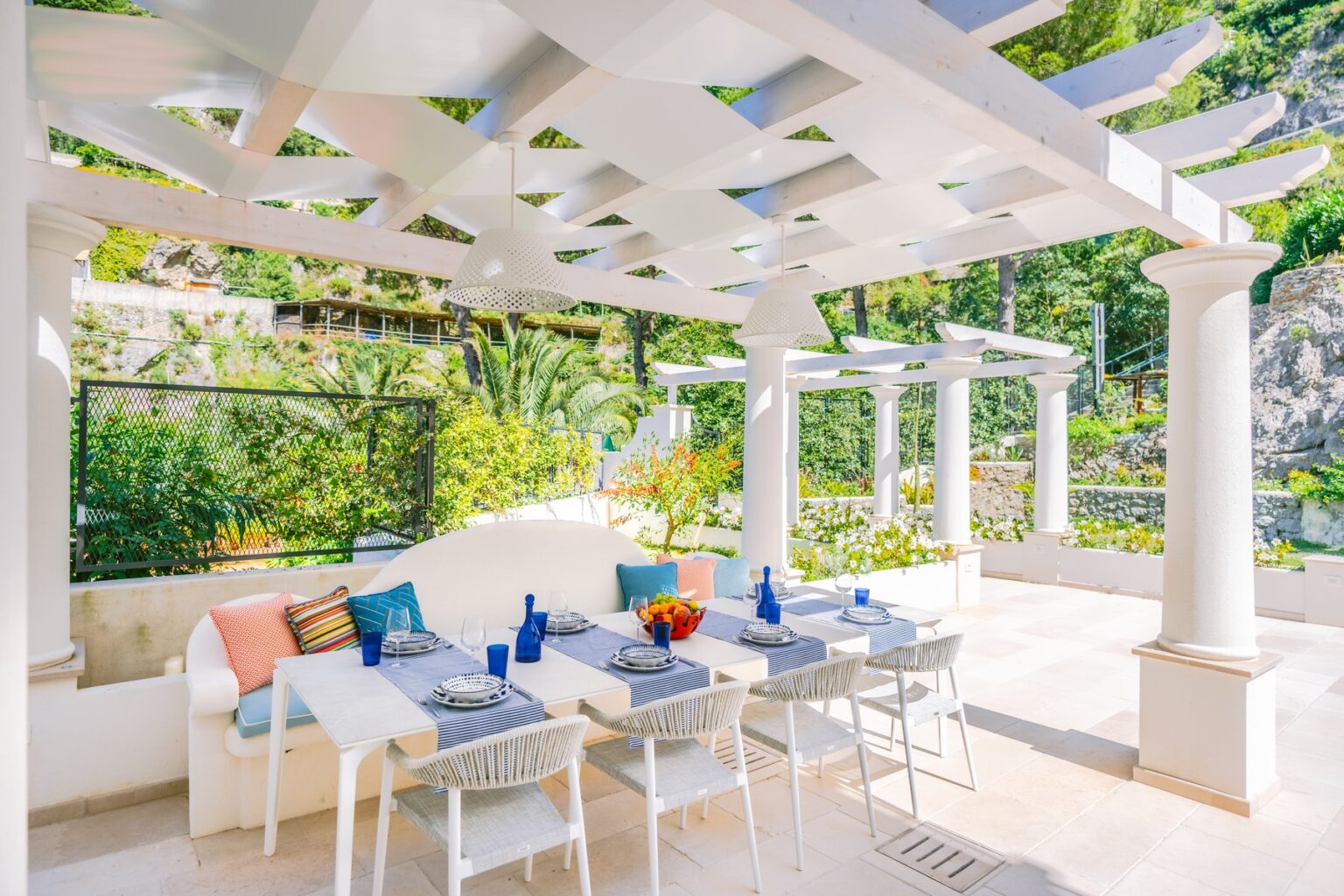 Elegant outdoor dining area featuring white columns, a trellis overhead, and stylish furniture. The space is adorned with colorful cushions and blue decor, surrounded by lush greenery, perfect for al fresco meals. | Only on SopranoVillas