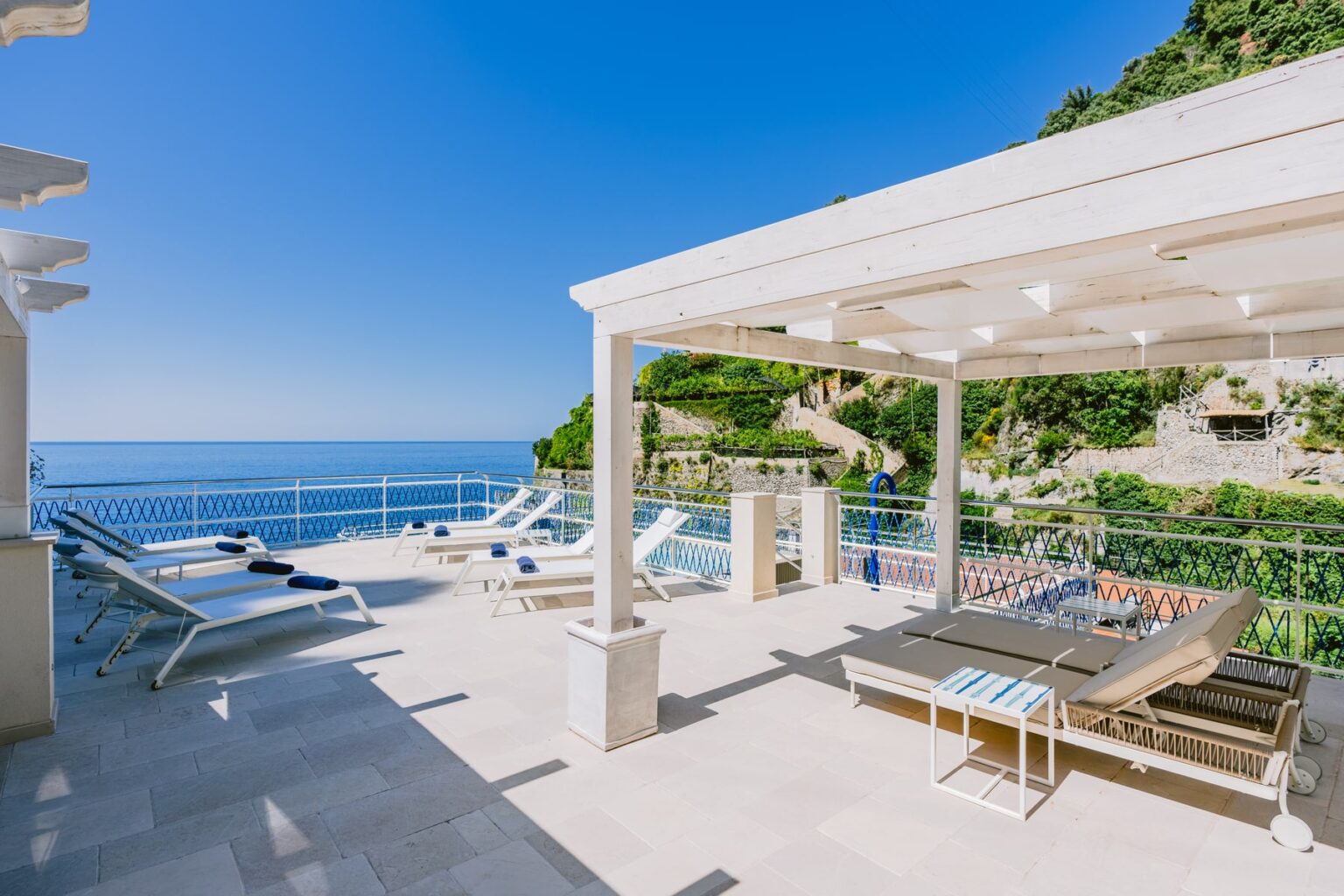 Luxury villa terrace overlooking the ocean with sun loungers and a swimming pool, surrounded by lush greenery and clear blue skies. Perfect for relaxation and outdoor enjoyment. | Only on SopranoVillas