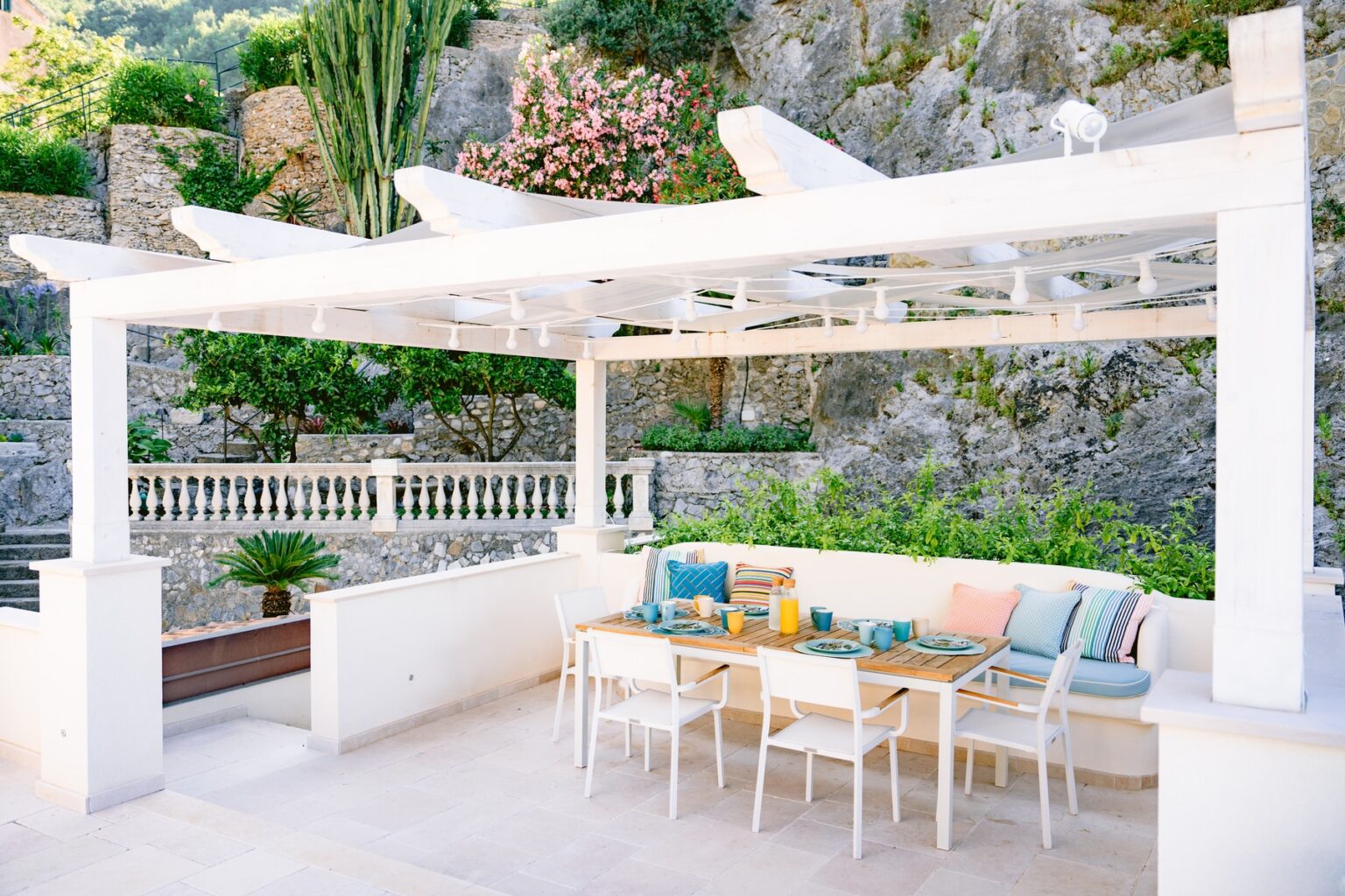 Outdoor dining area with a white pergola, modern wooden table, and colorful cushions, surrounded by lush greenery and stone walls. Ideal for al fresco meals and relaxation in a serene garden setting. | Only on SopranoVillas